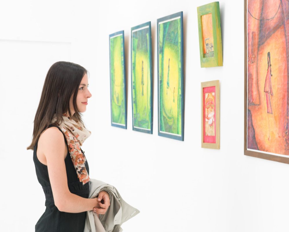 side view of young caucasian woman standing in an art gallery in front of colorful framed paintings displayed on a white wall